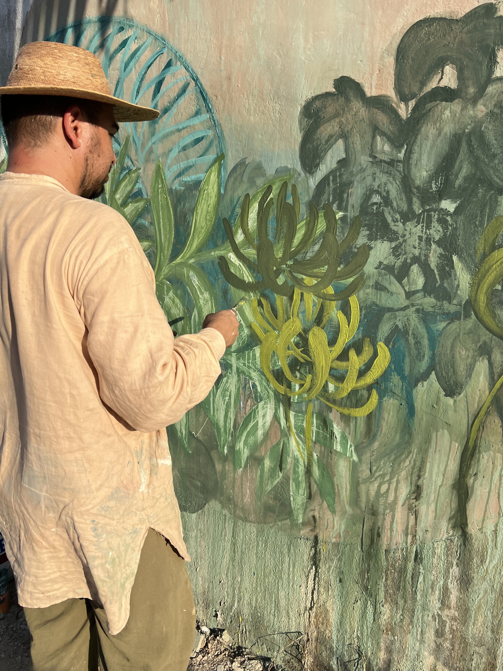 Artista con sombrero pinta hojas de selva en un mural verde