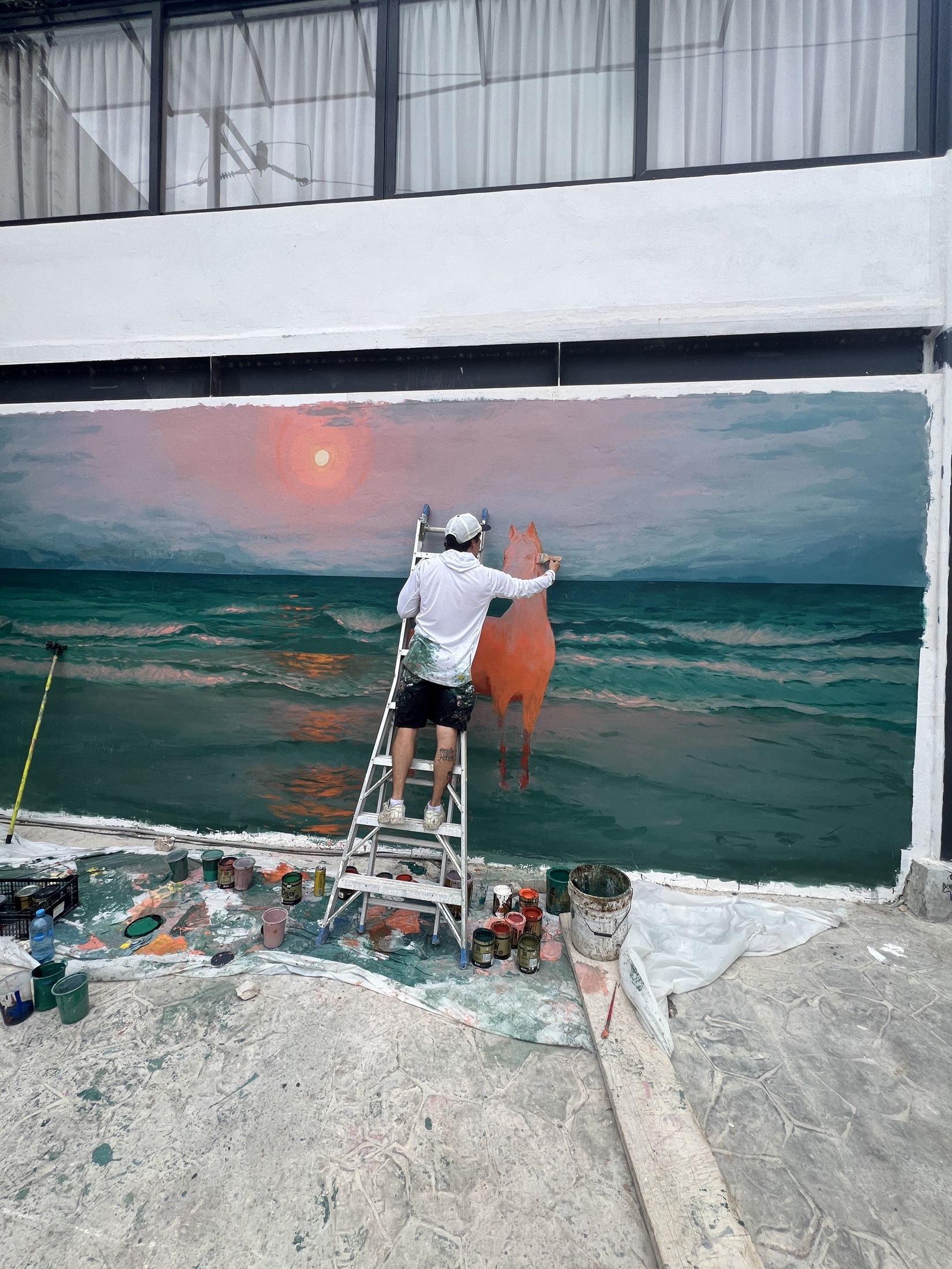 Vista completa del mural del mar con el artista en la escalera y botes de pintura en el piso